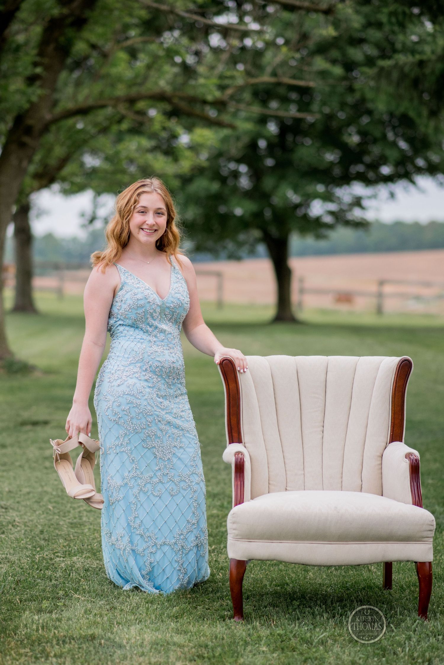 Woman in a sparkling blue gown stands beside a cream-colored vintage armchair on manicured grounds.