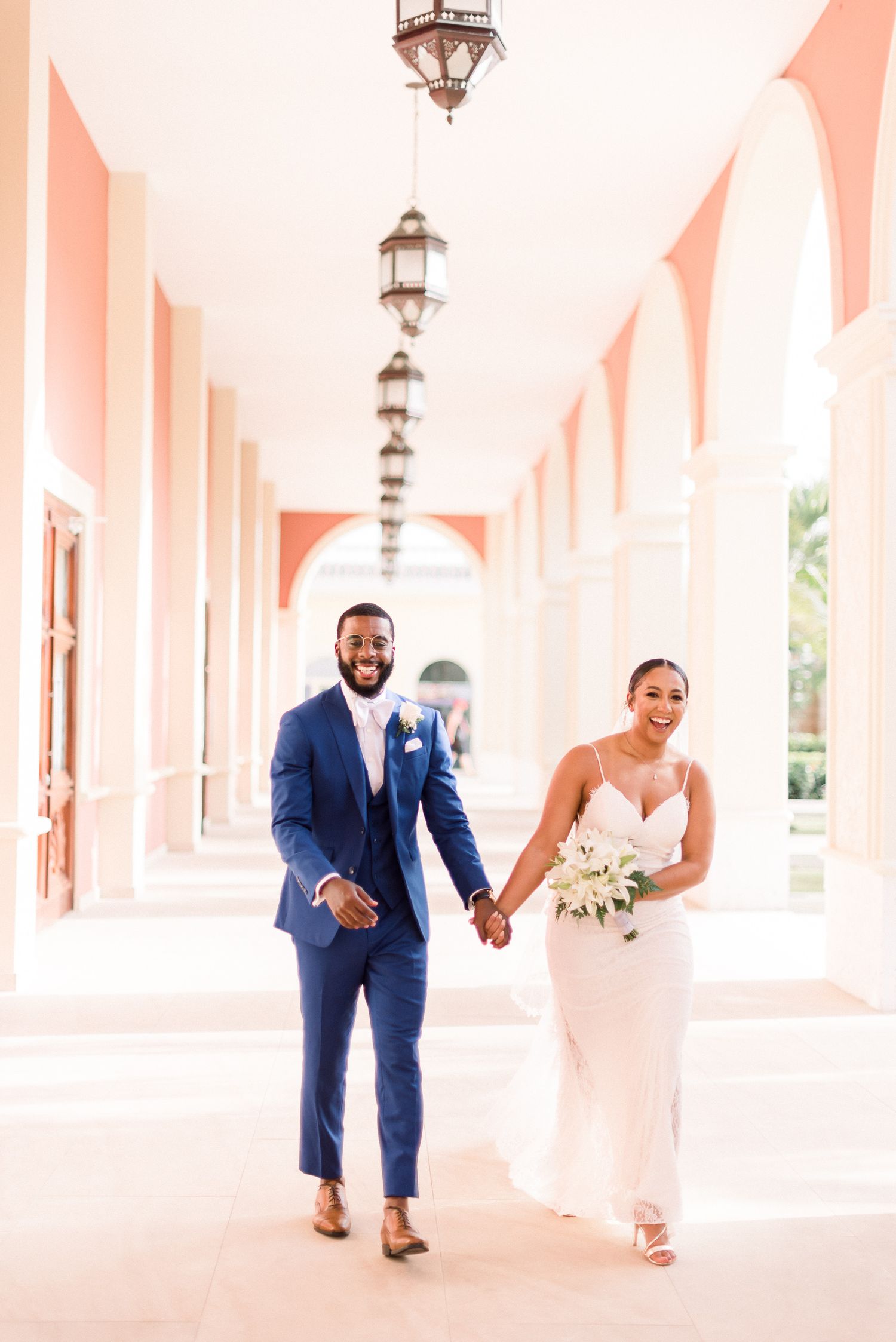 A wedding couple walking hand in hand down an elegant coral-colored colonnade hallway.