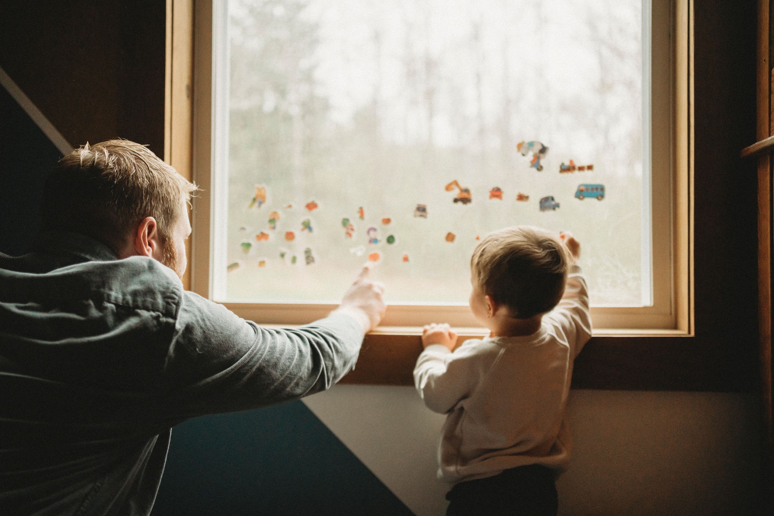A parent and child look out a window decorated with colorful stickers as sunlight streams in.