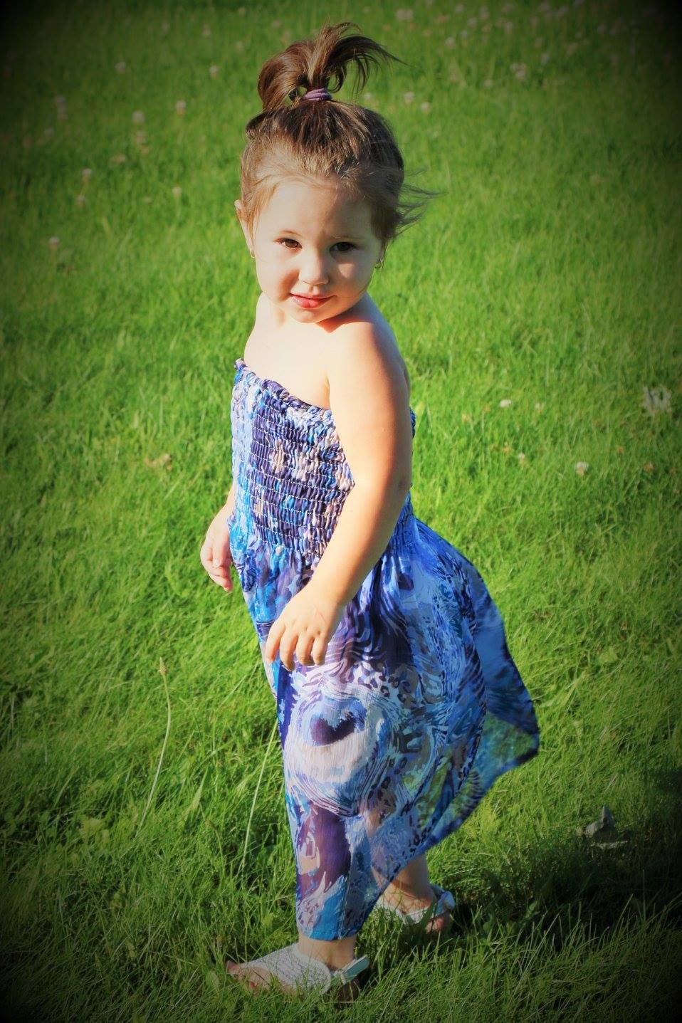 A small child wearing a blue tie-dye sundress twirls playfully on green grass.