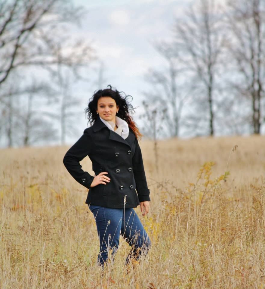A person in a black peacoat stands confidently in a autumn field with bare trees in background.