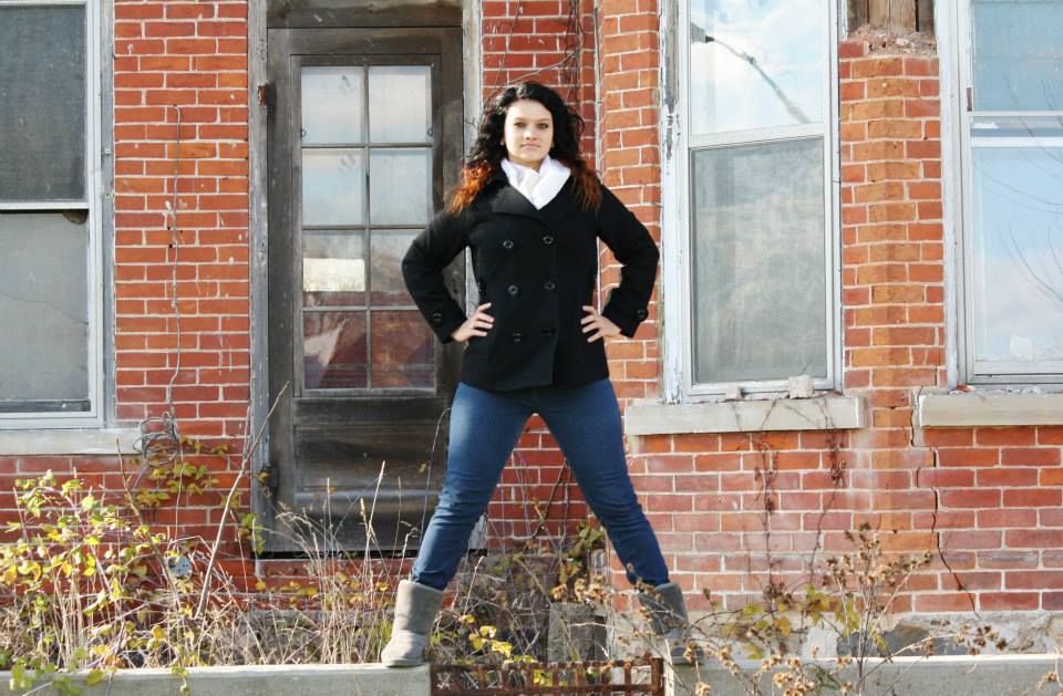 A person strikes a confident pose in black jacket and jeans against a brick building.