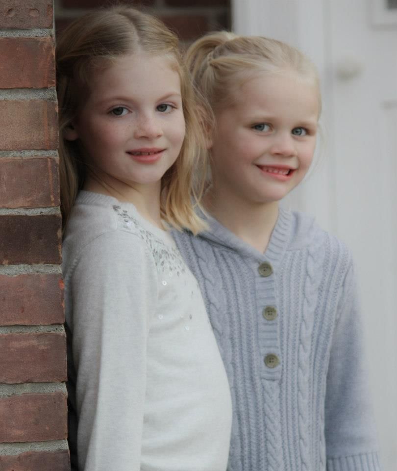 Two children wearing white and gray sweaters pose together against a brick wall.