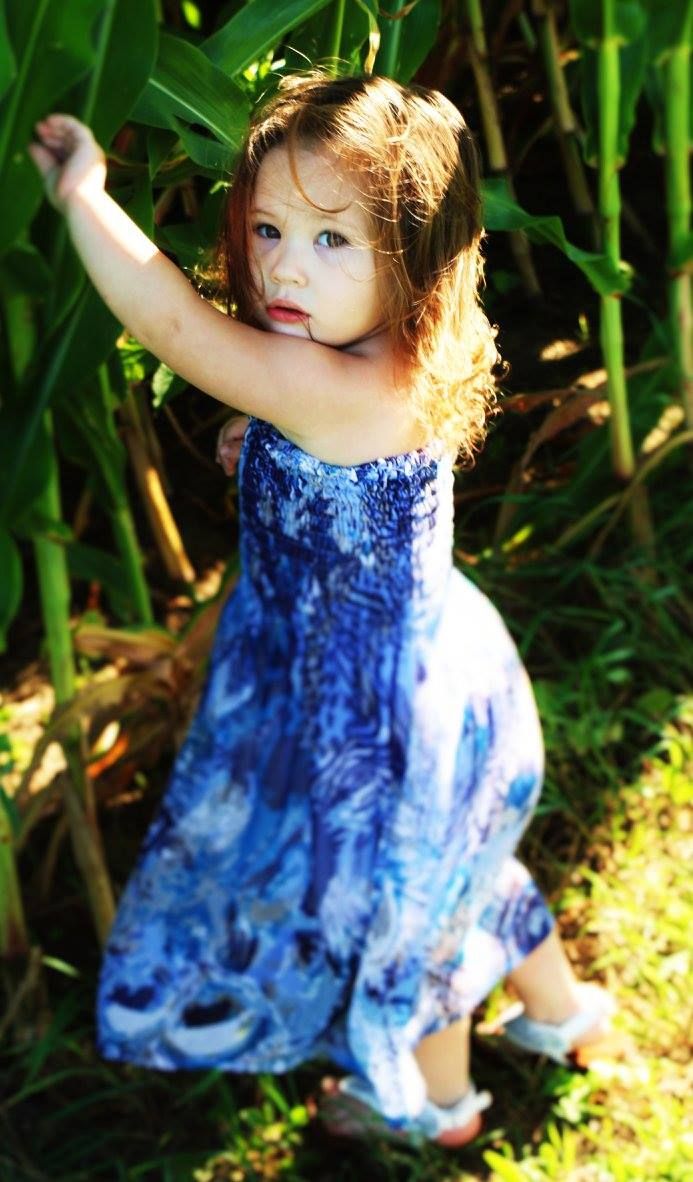 A child in a flowing blue summer dress poses among tall green plants in dappled sunlight.