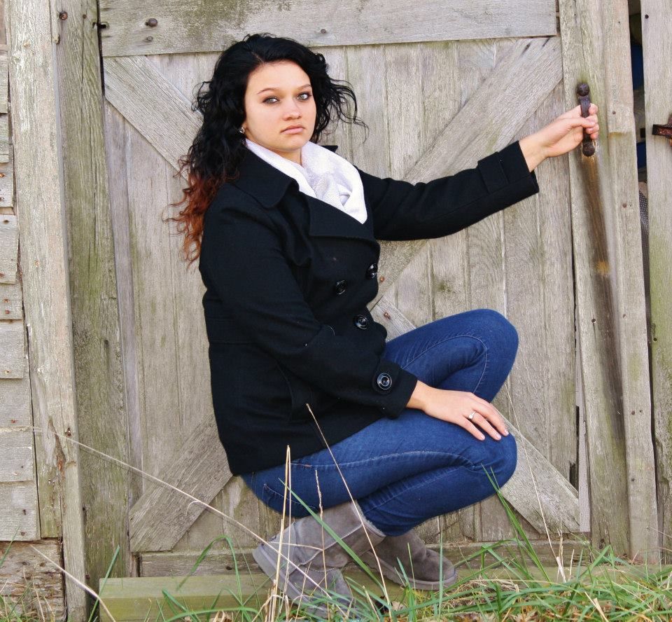 A person in black jacket and jeans poses casually against a rustic wooden door.