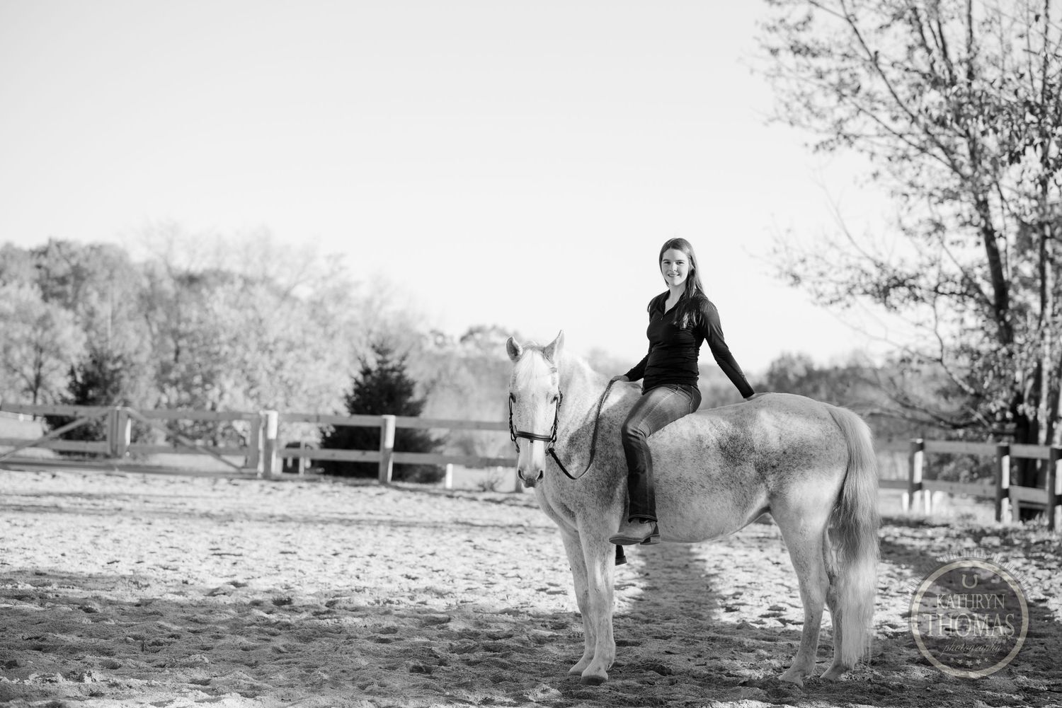 Black and white image of person riding white horse in sandy arena.