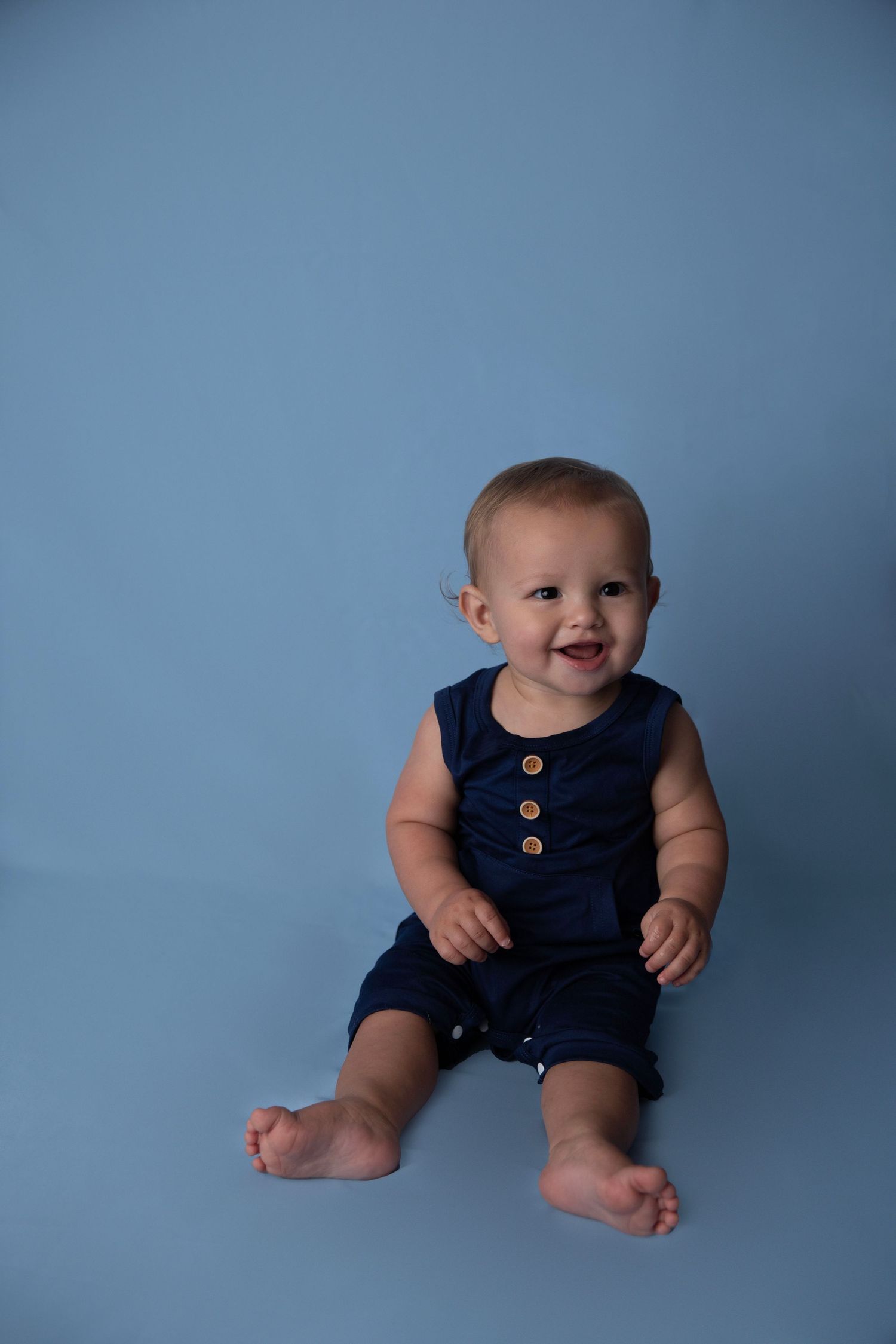 Cheerful baby boy in navy blue romper sitting barefoot against a matching blue background.