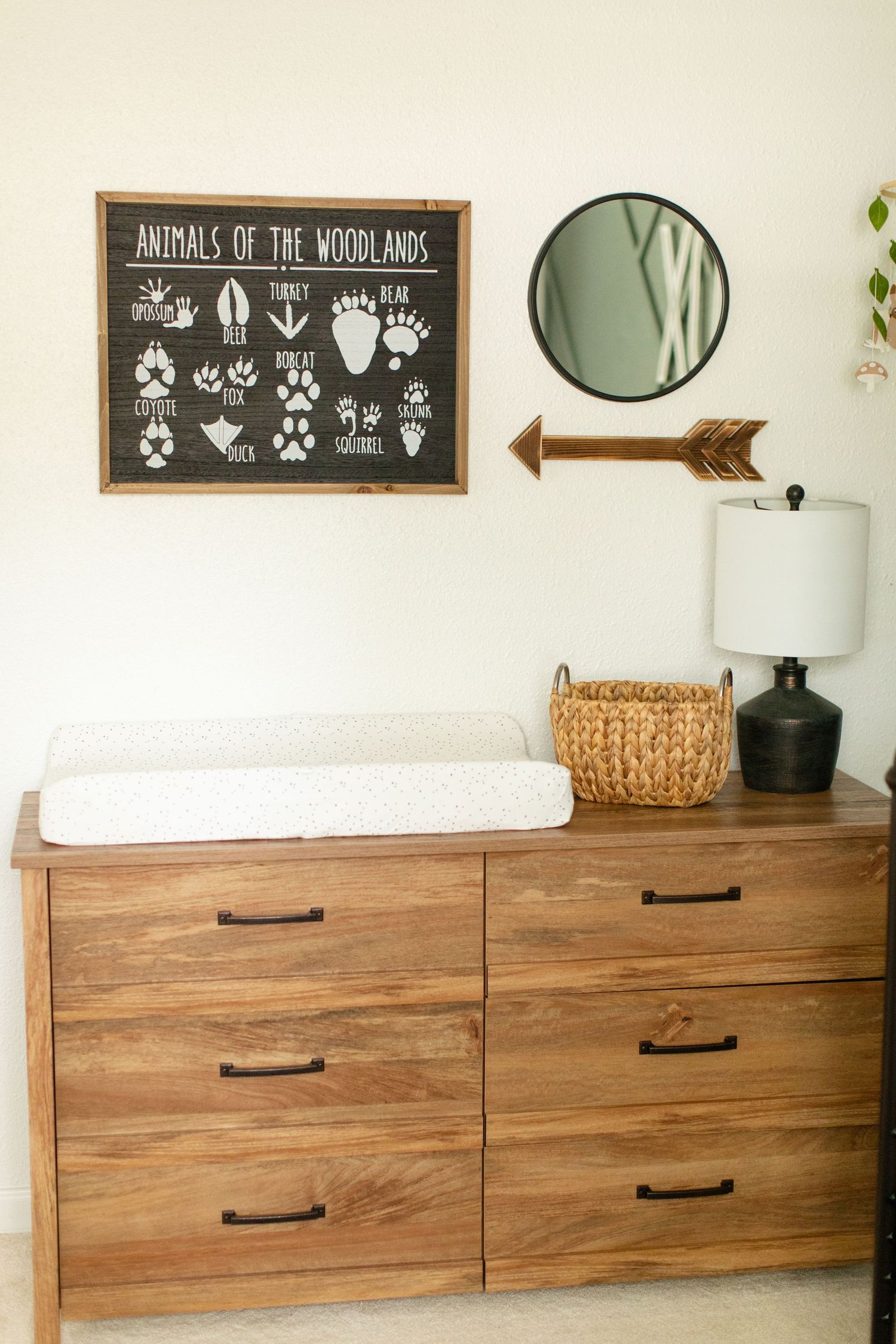 A nursery dresser with changing pad features a woodland animal sign, round mirror, and decorative arrow on white walls.
