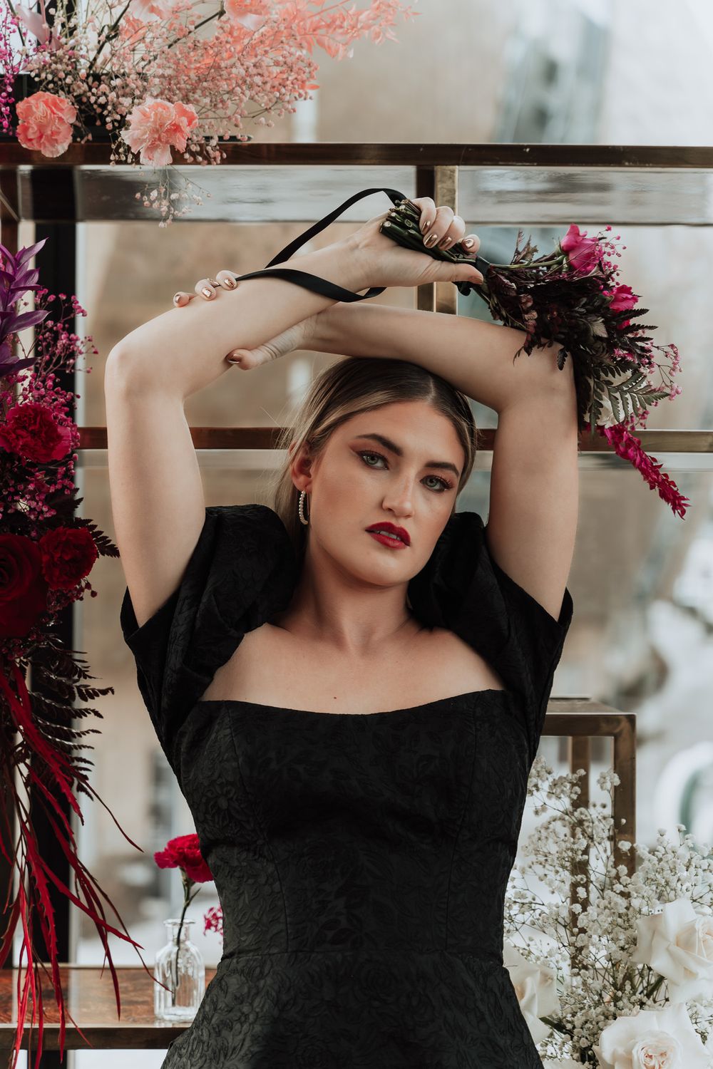 Dramatic pose in black formal dress with raised arms framed by pink and red florals.