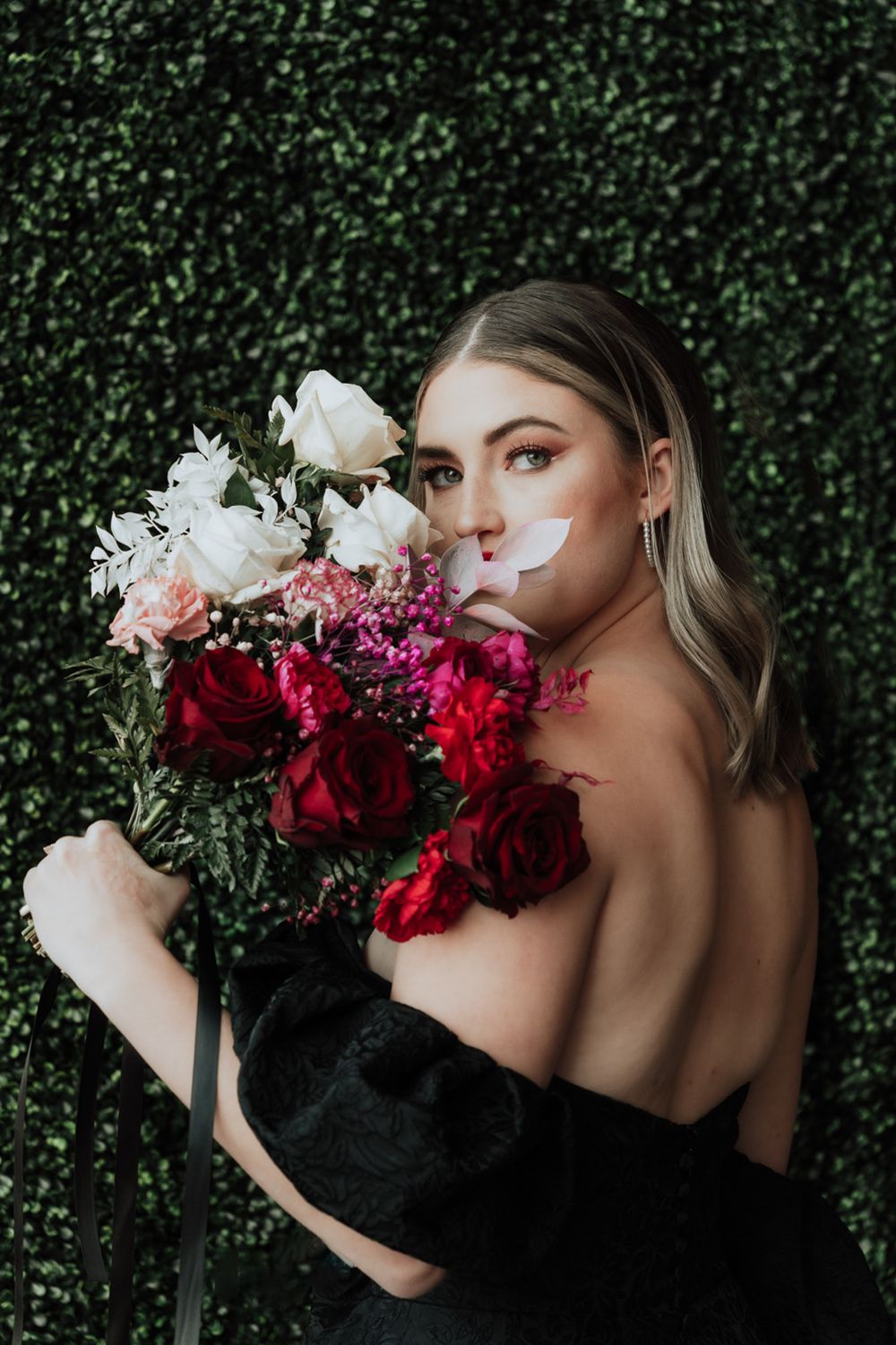Elegant portrait with roses featuring model in black dress against green hedge backdrop.