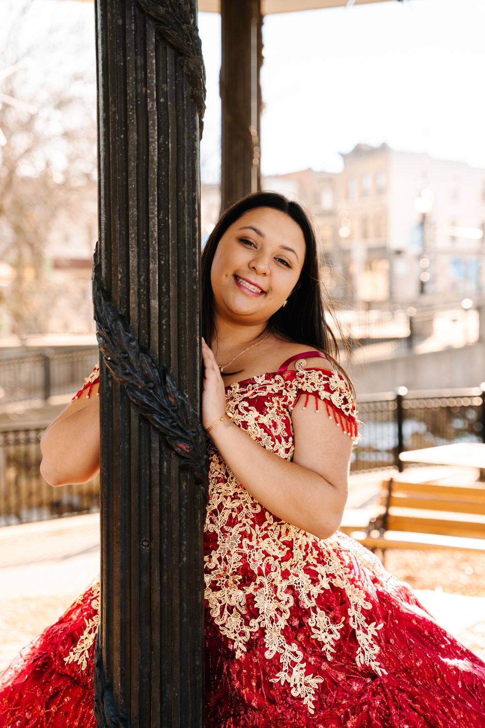 Person in red and white embroidered ballgown poses by vintage black column on sunny porch.