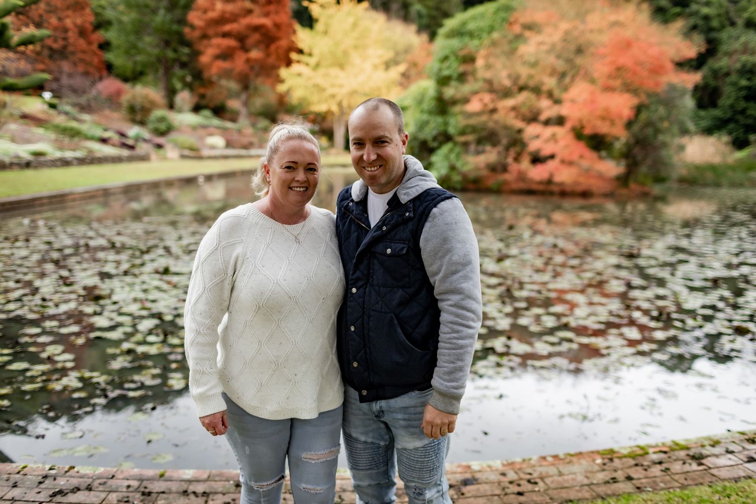 A couple poses together by a pond with autumn trees creating a colorful backdrop.