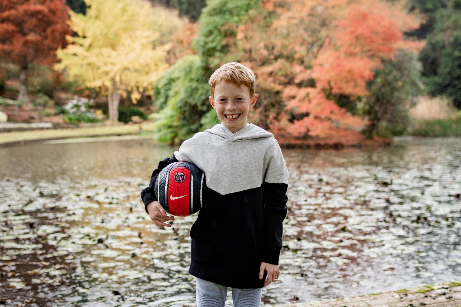 A person holding a red and black soccer ball stands by a pond with vibrant autumn foliage in the background.
