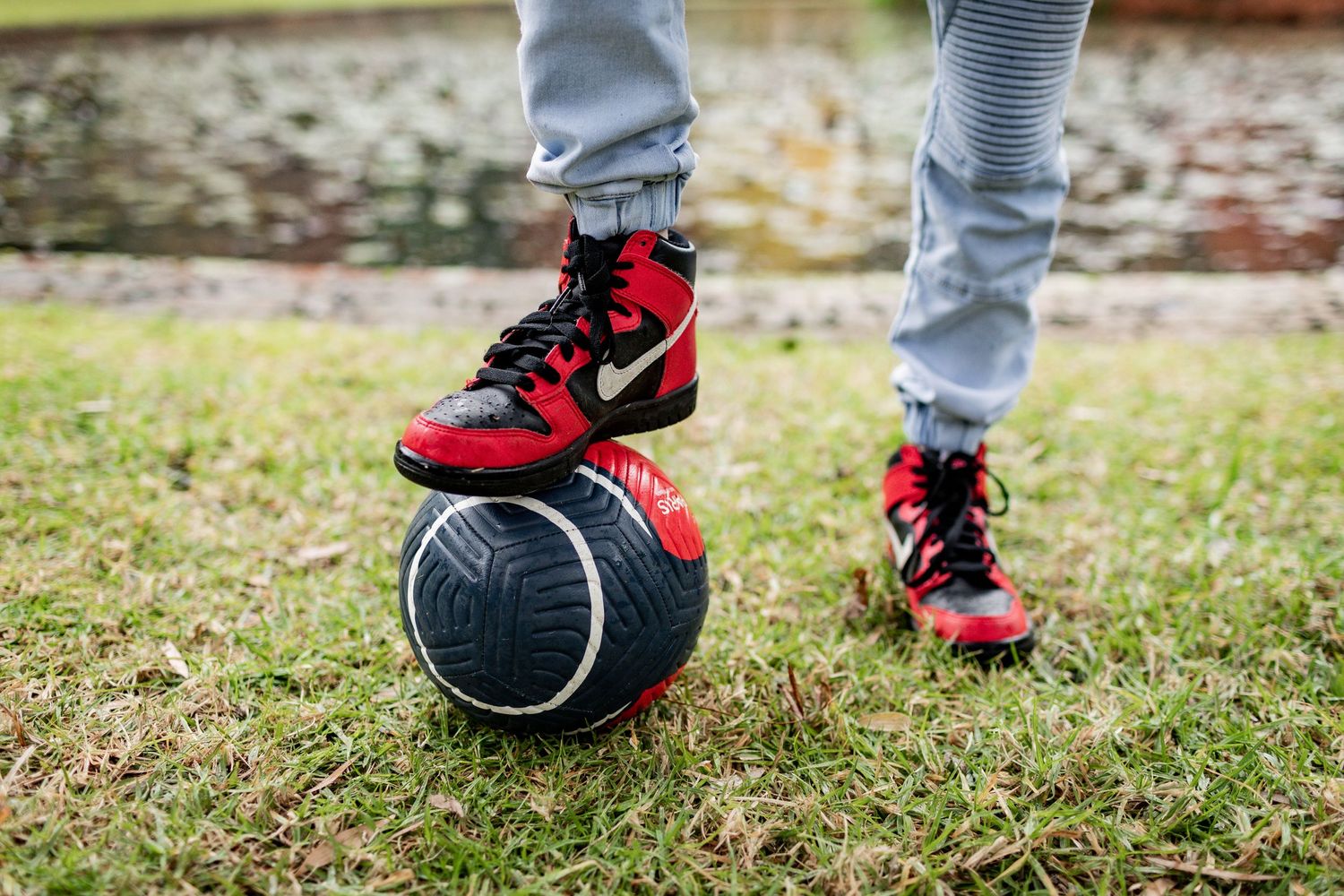 Red and black Nike sneakers resting on a soccer ball against grass and autumn leaves.