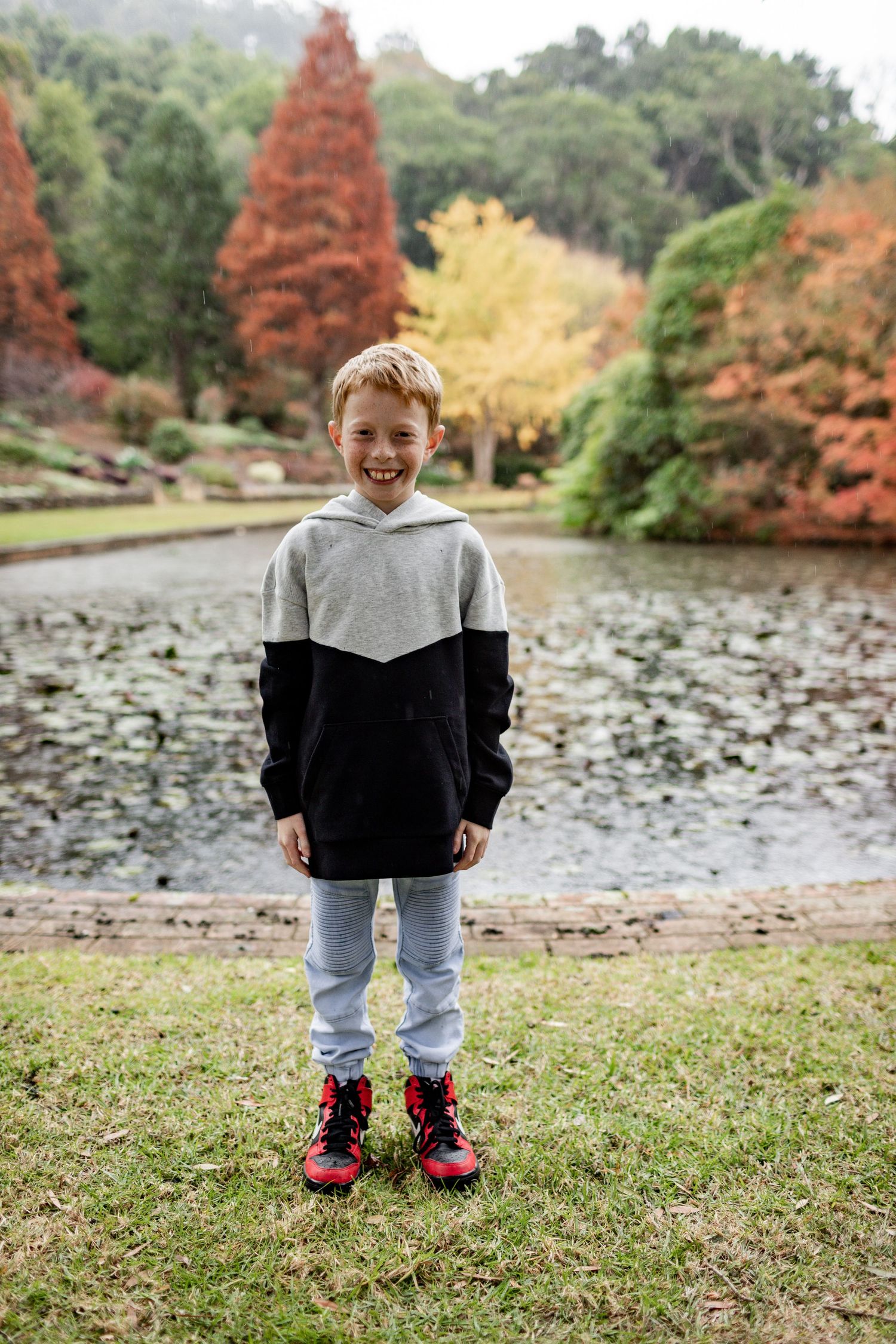 Person in black and gray hoodie standing by pond with fall foliage in background.