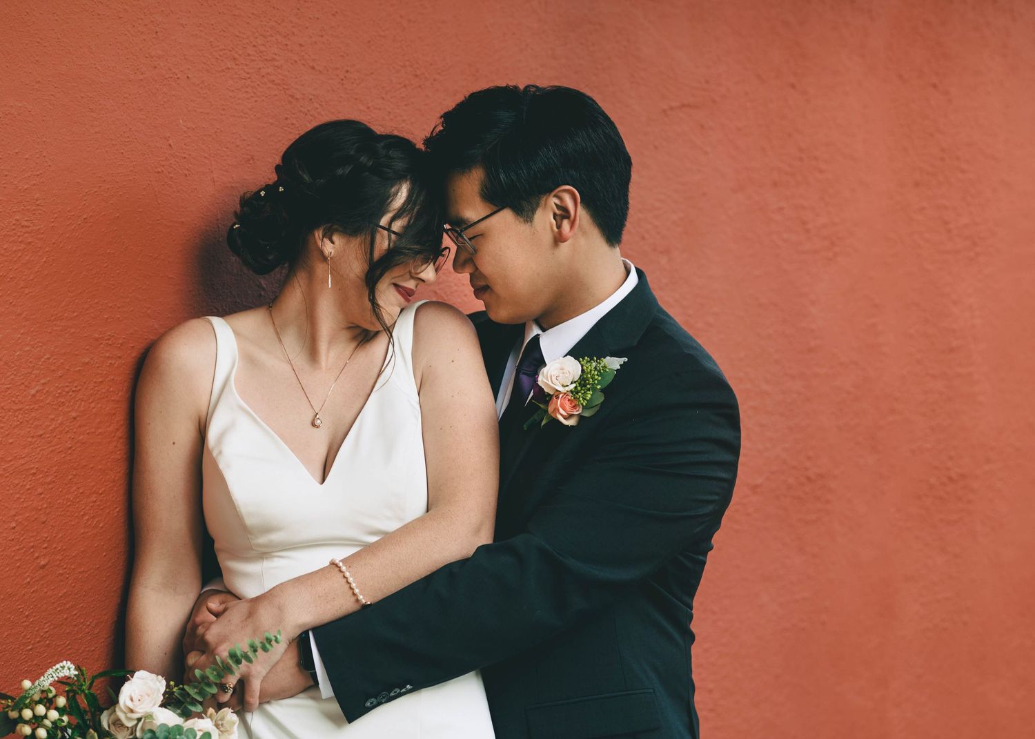 Bride and groom, heads touching, in front of the red Adobe walls at Cooper Molera in Monterey.
