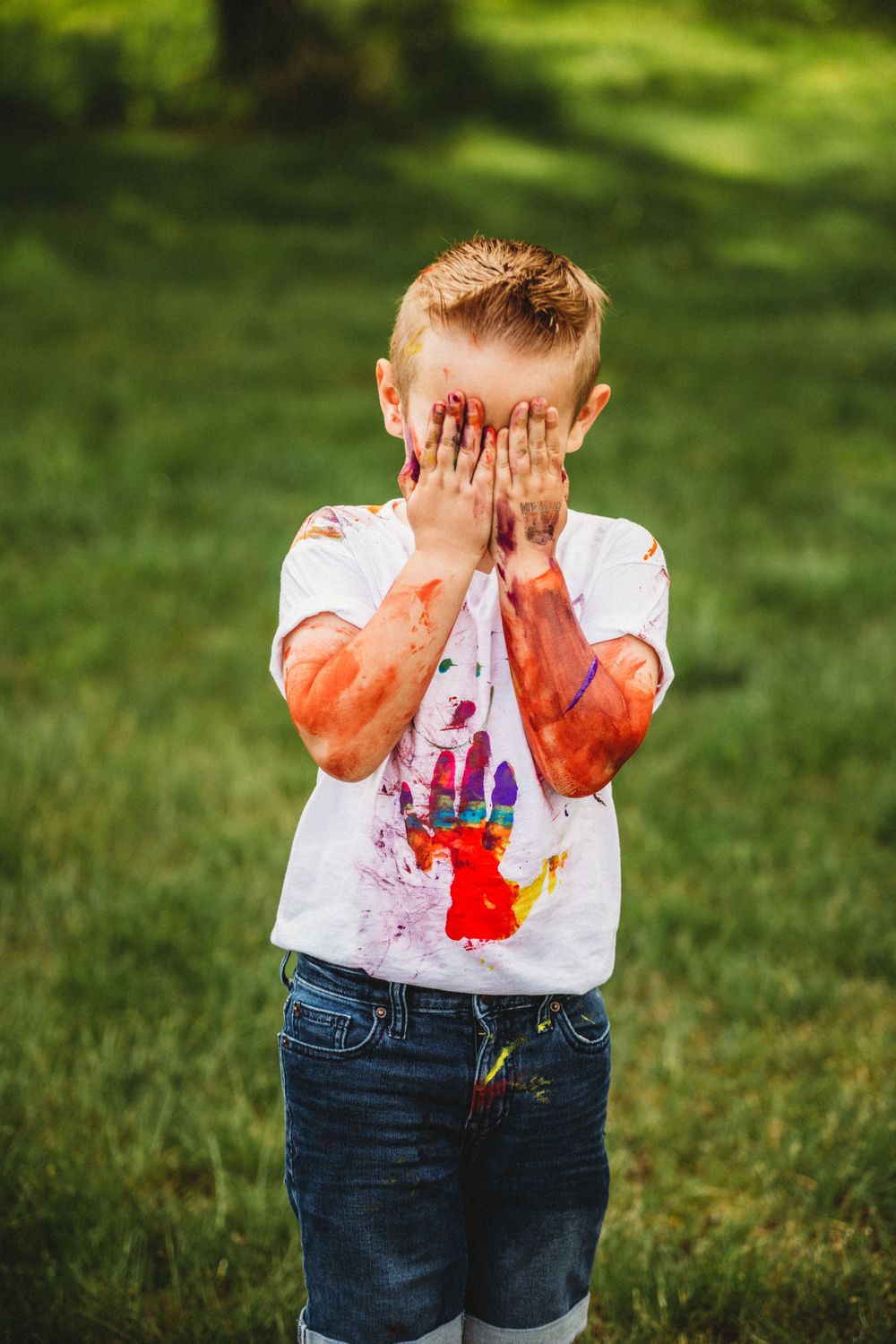 Child in white shirt and denim shorts with red paint covering hands and face.