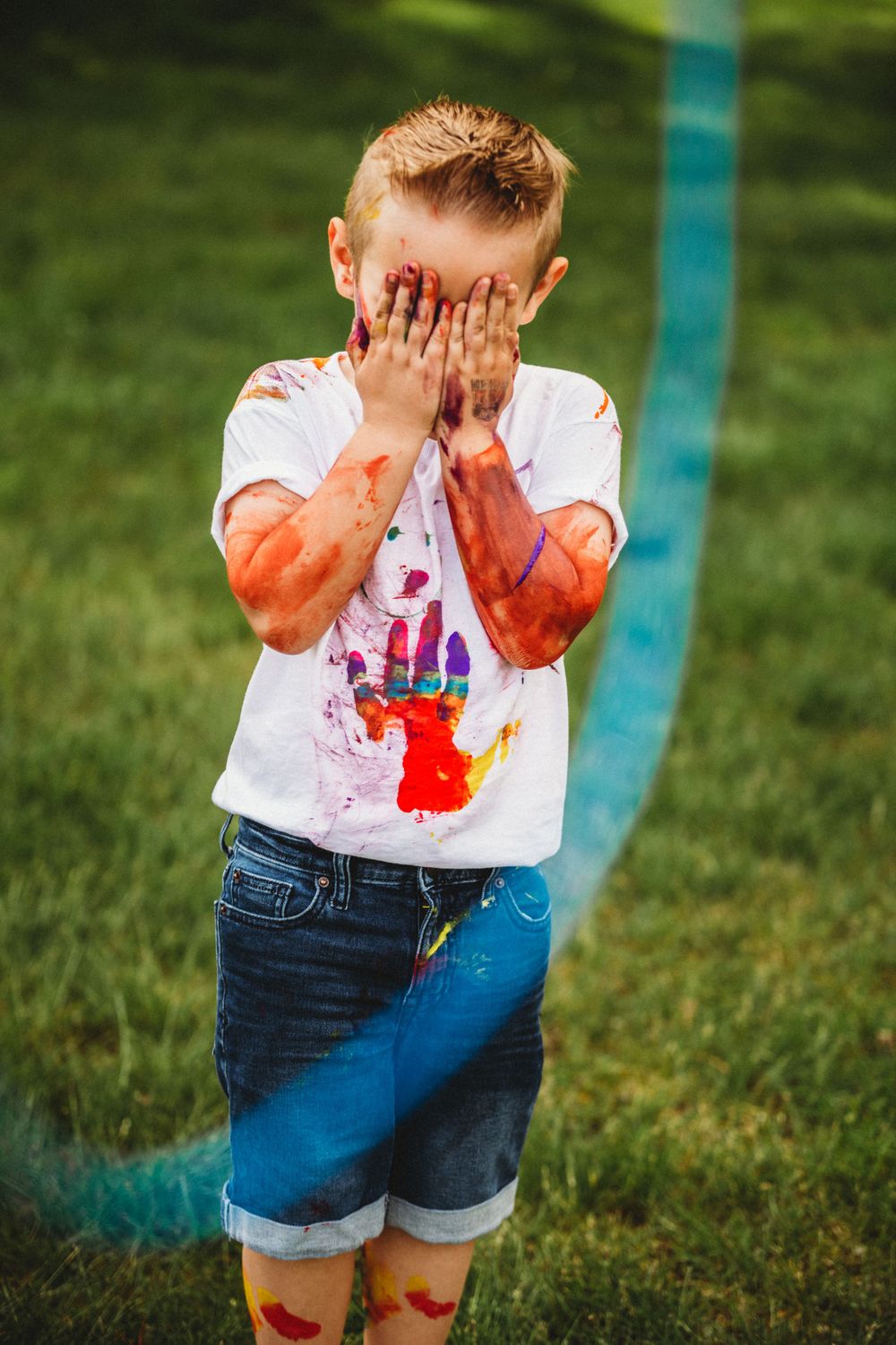 Child with painted hands covering face while standing on grass.