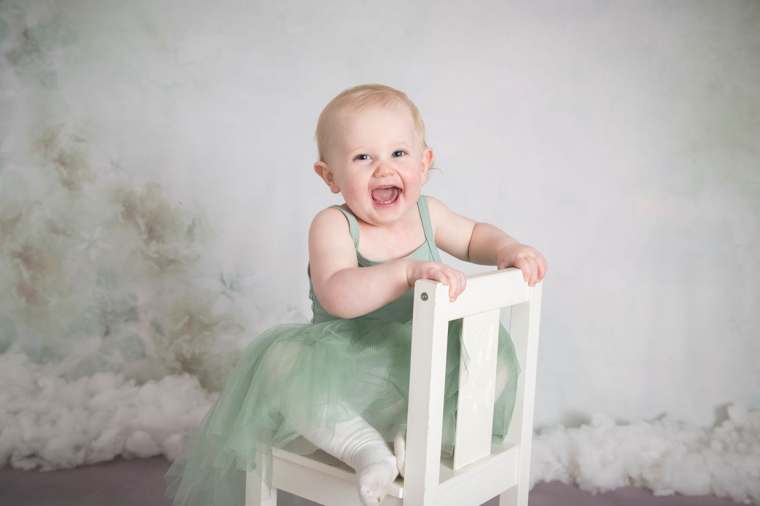 Baby in mint green tutu dress standing on white chair against grey backdrop.