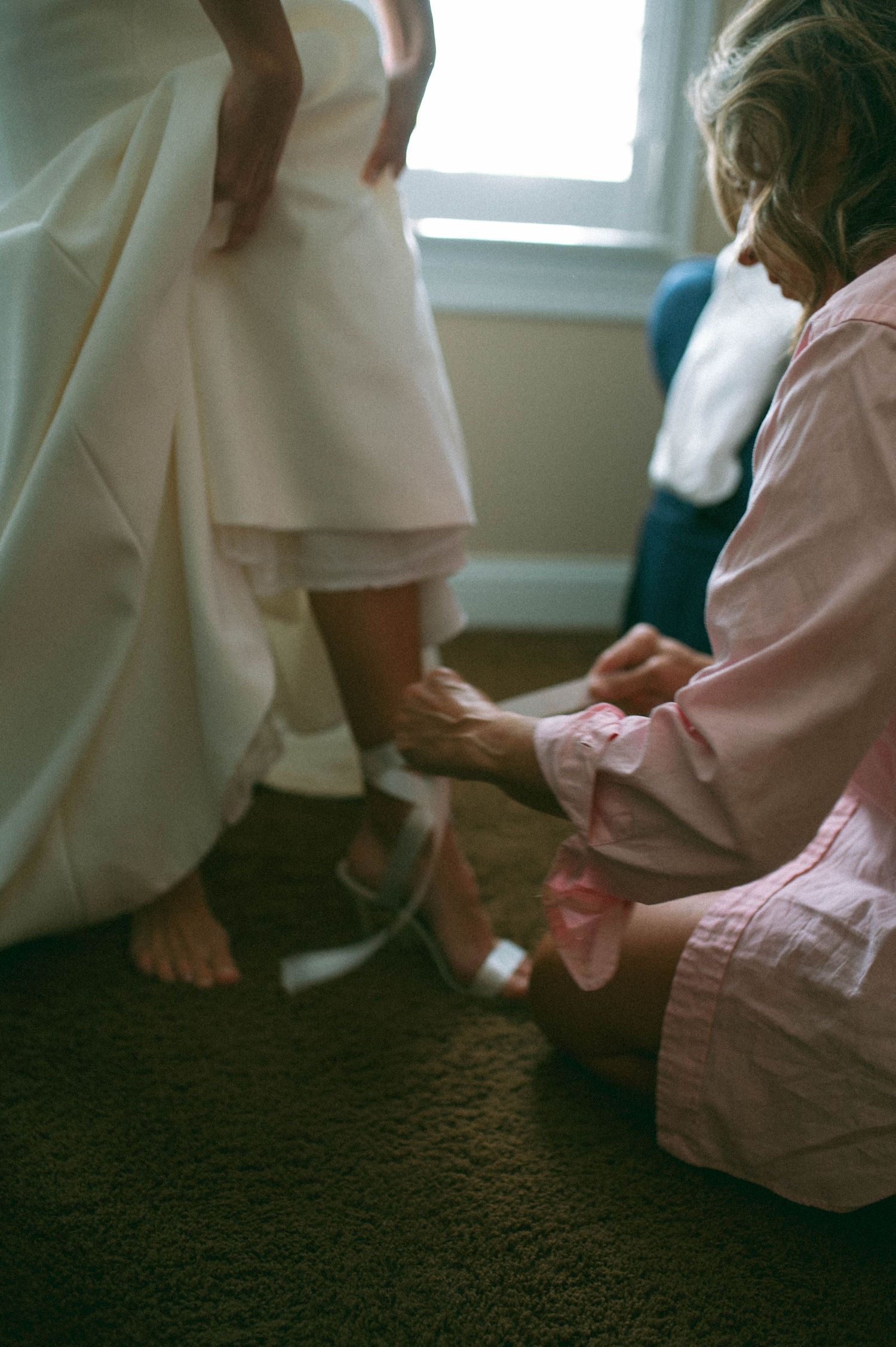 A person in pink helps dress a bride in her white wedding gown as they get ready in a room.