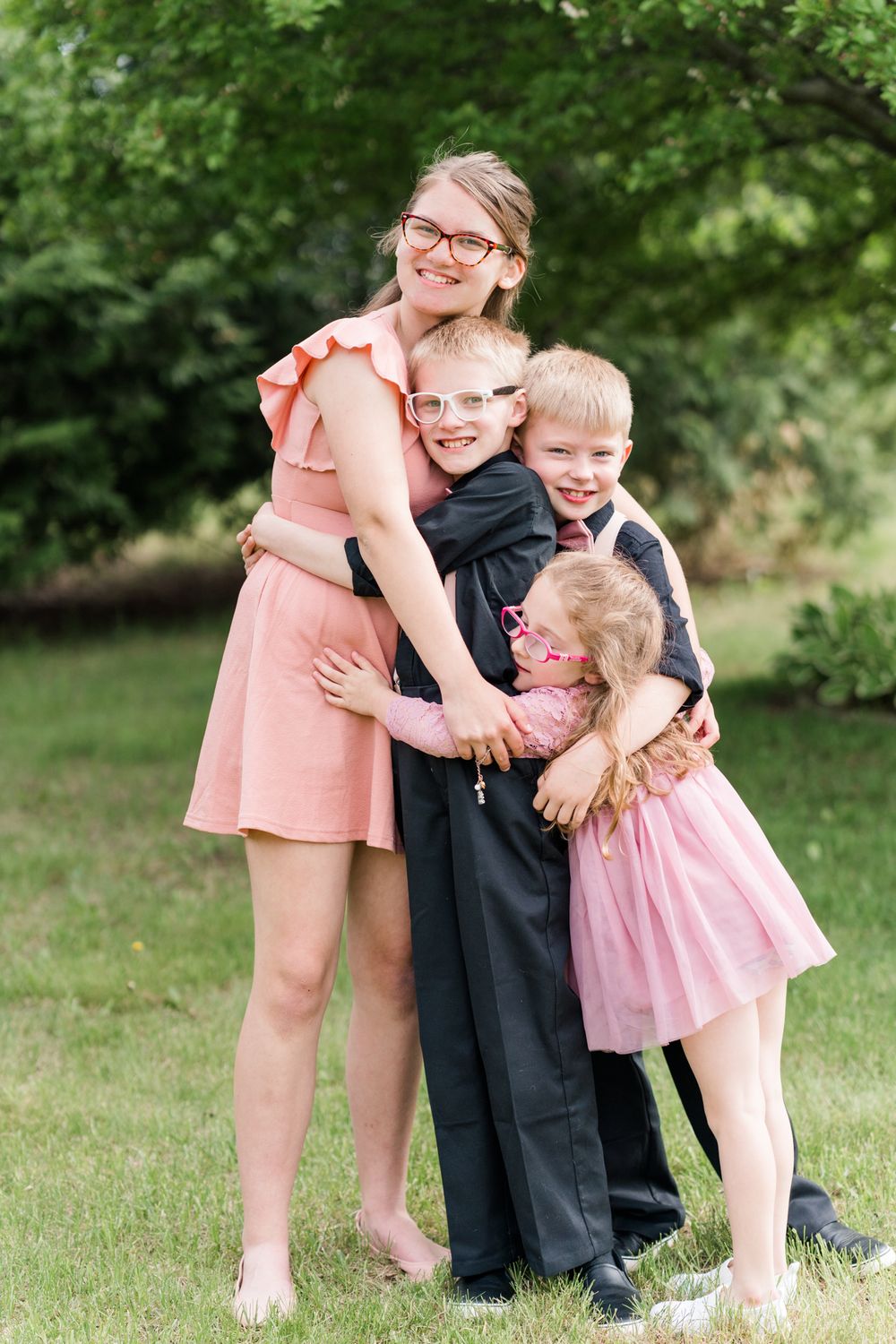 Three siblings dressed in formal attire share a joyful group hug outdoors in a garden setting.