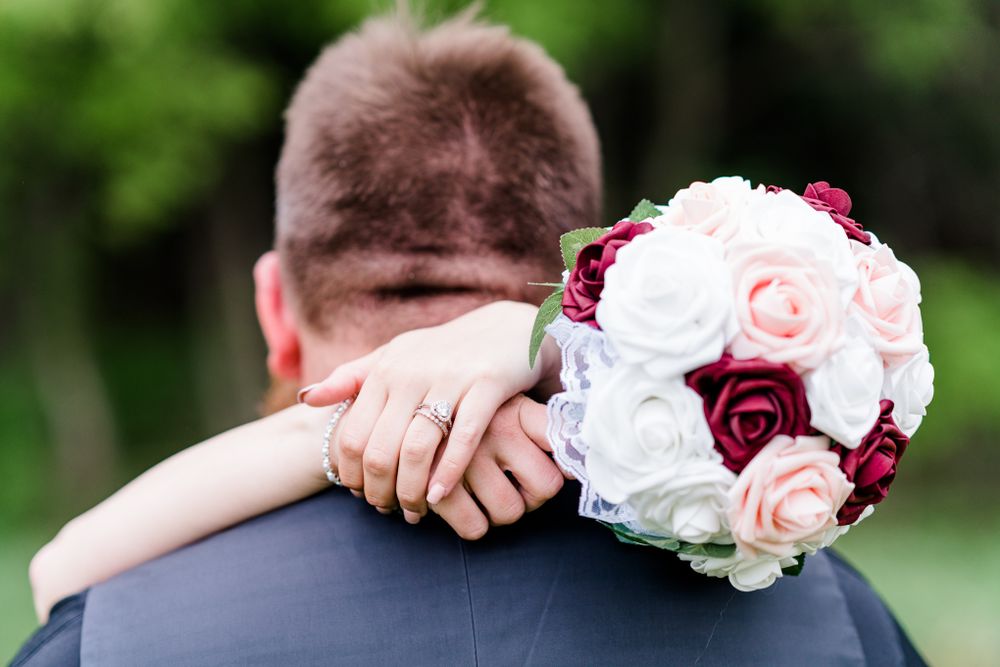 A wedding bouquet with white and burgundy roses rests between embracing newlyweds.