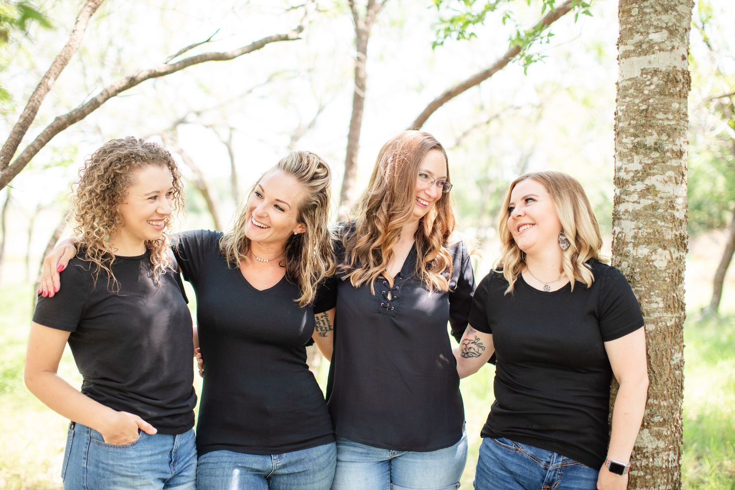 Four friends in matching black shirts laugh together outdoors in natural lighting.