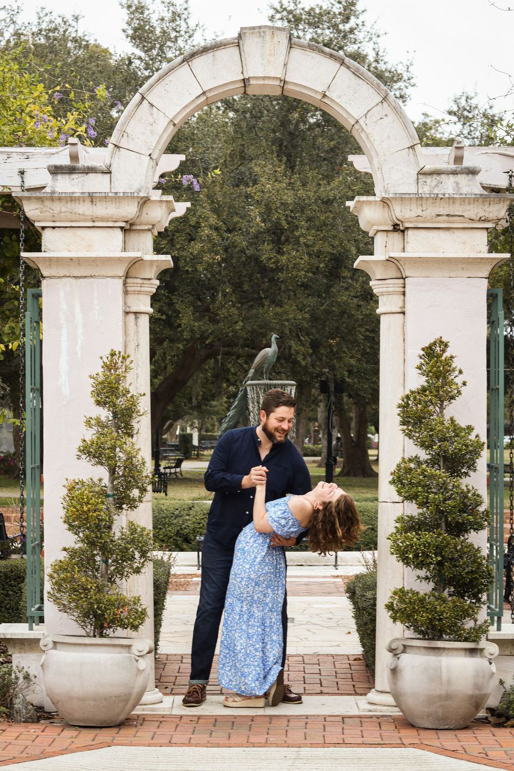 A romantic dip pose under a white stone archway with potted plants.