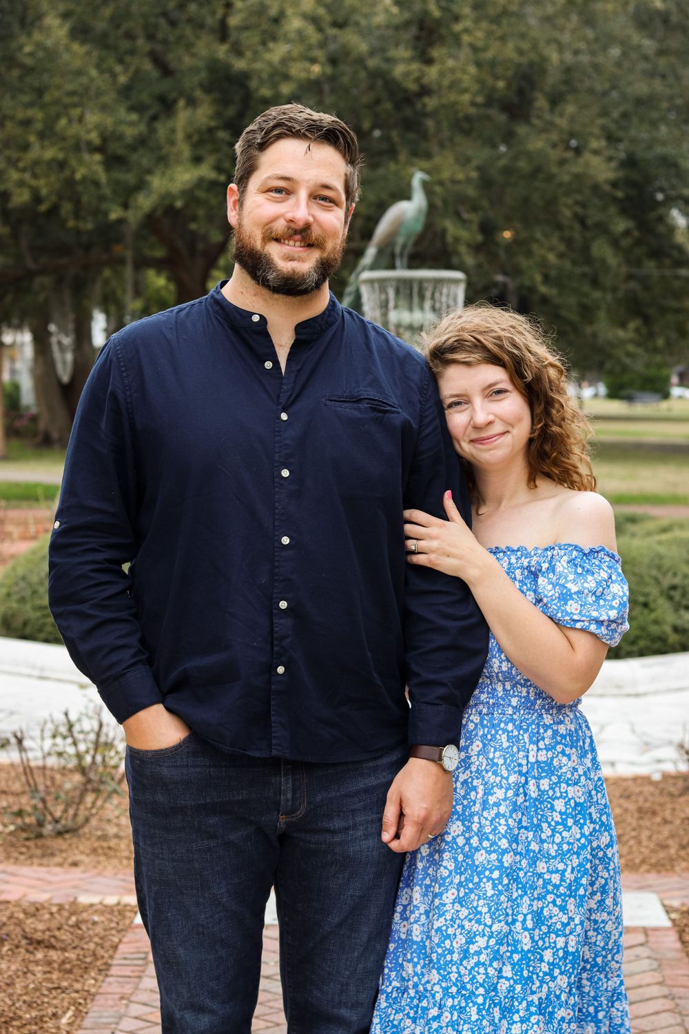 A couple in navy and blue outfits pose together outdoors near garden features.