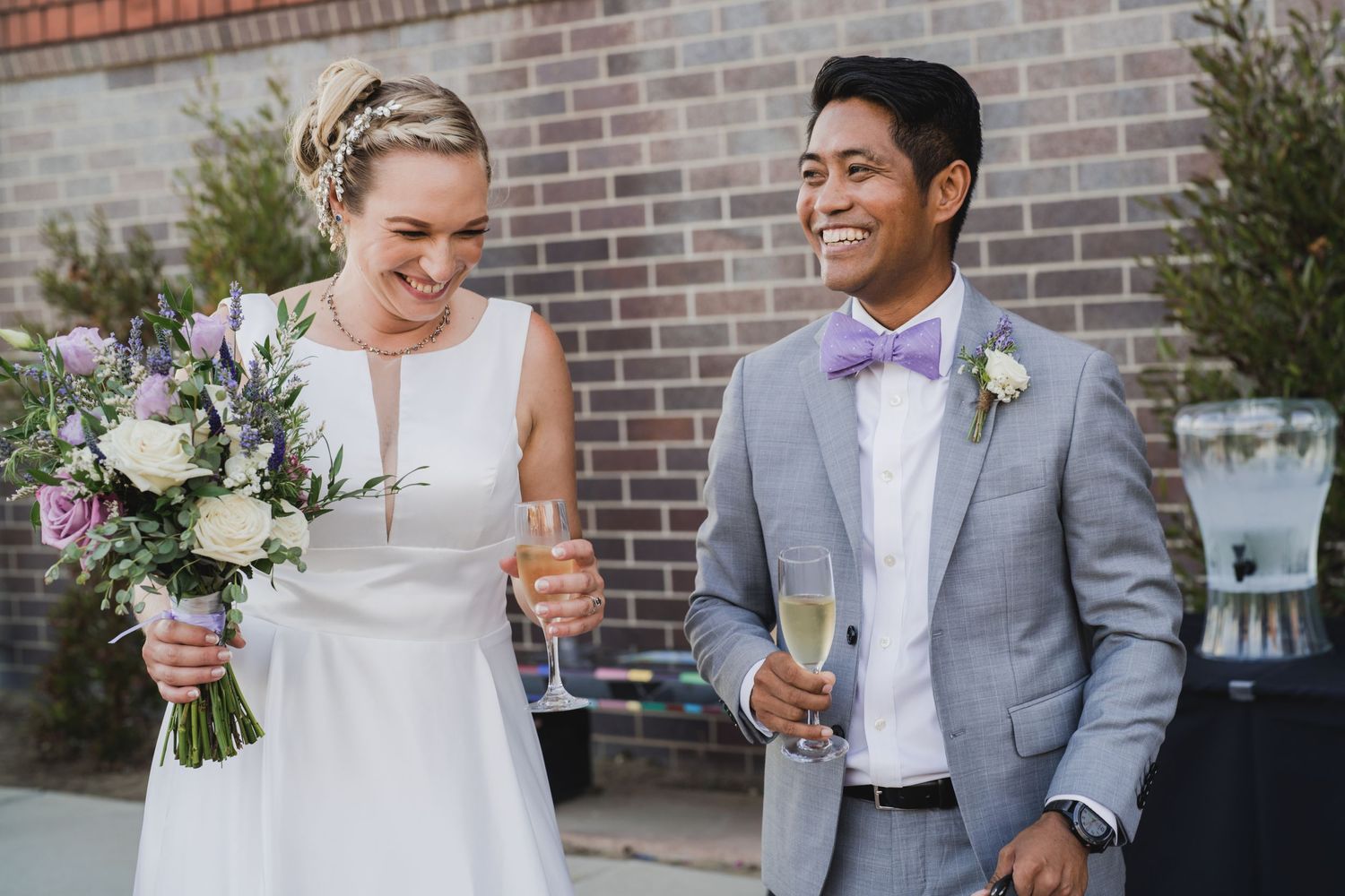 Wedding couple shares joyful moment against brick wall with bride holding floral bouquet.