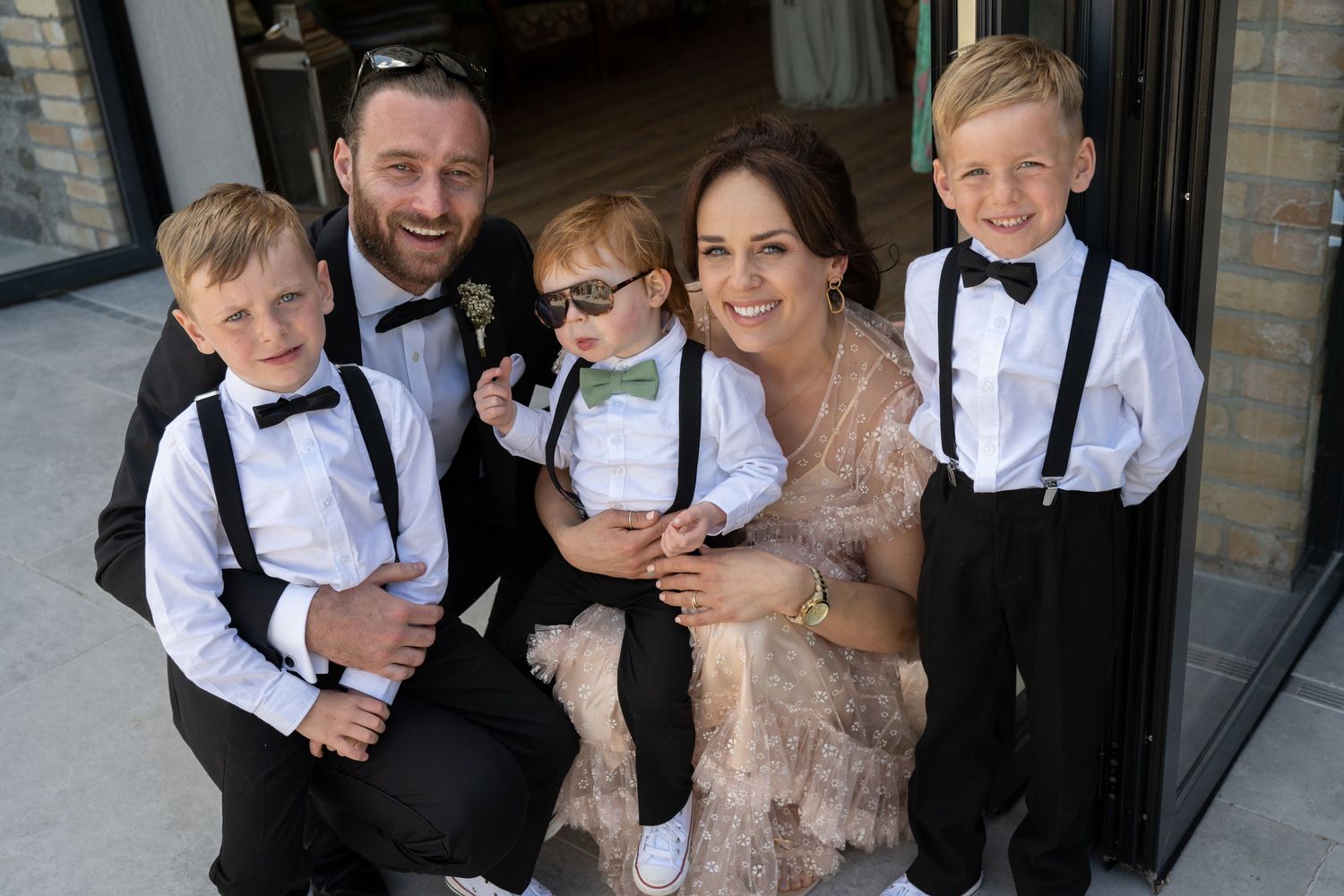 Family dressed in formal attire with children in bow ties and suspenders pose together.