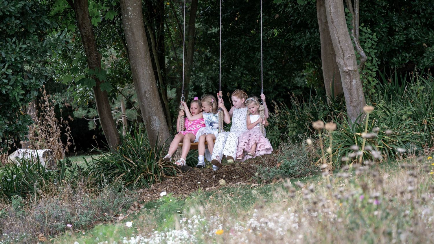 Children sitting together on a swing in a wooded garden area.