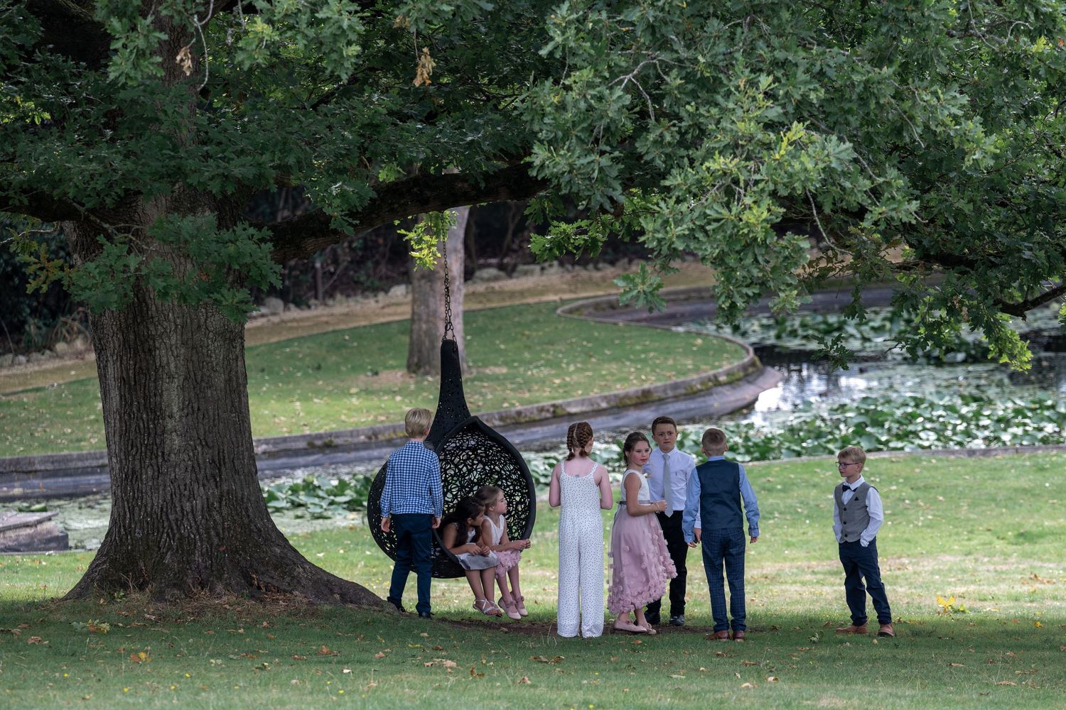 Wedding party posing for photos under a large tree in a scenic garden setting.