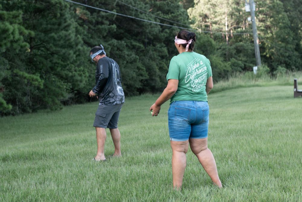 Two people walking through a green field with pine trees in the background.