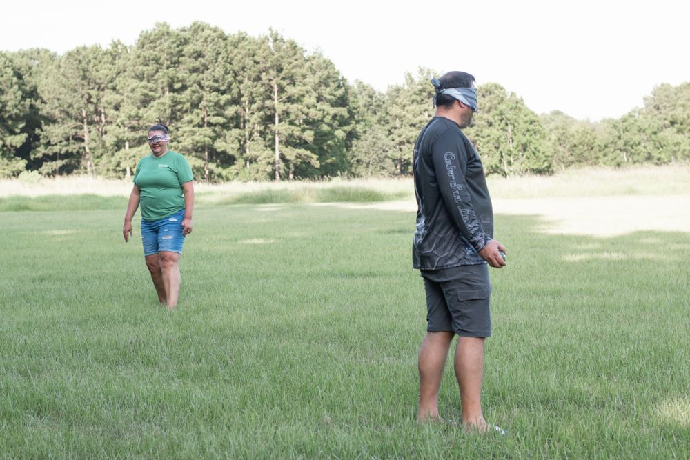 Two people standing in a grassy field surrounded by trees on a sunny day.