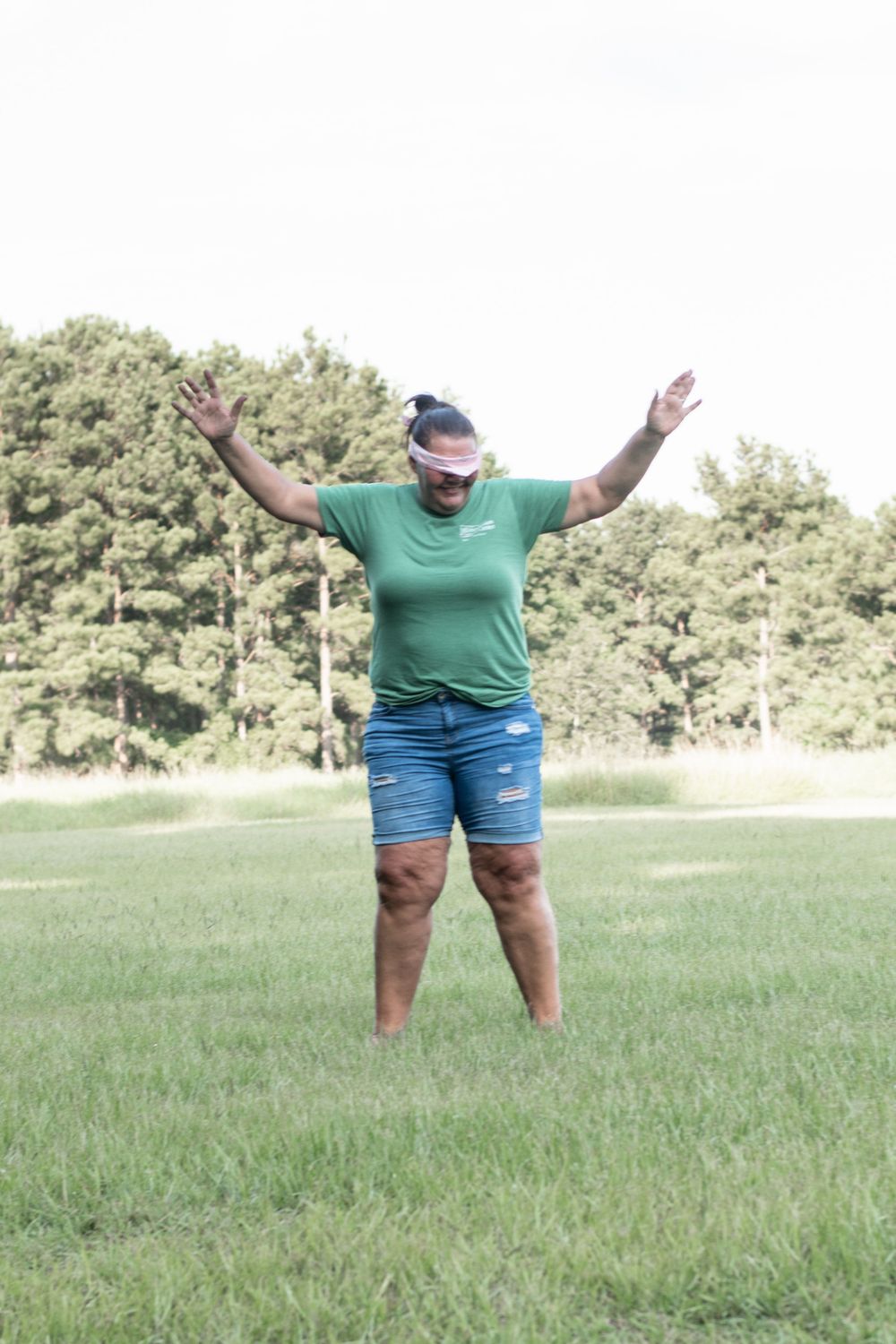 Person in green shirt and denim shorts standing with arms outstretched in grassy field.