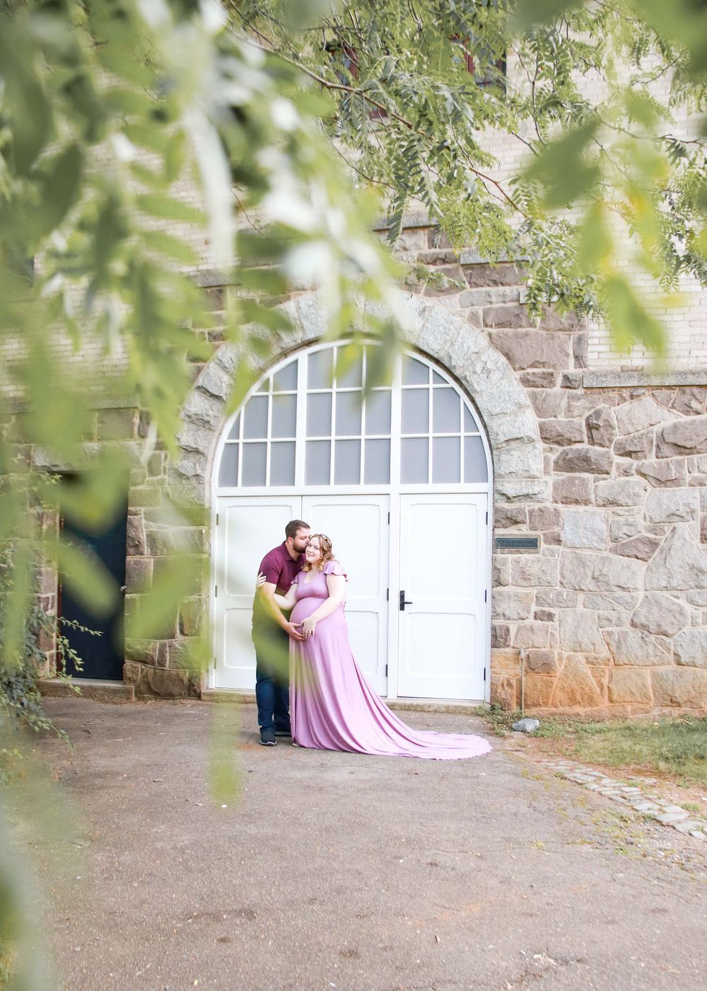 Romantic couple embraces in front of arched white door with stone wall and green foliage.