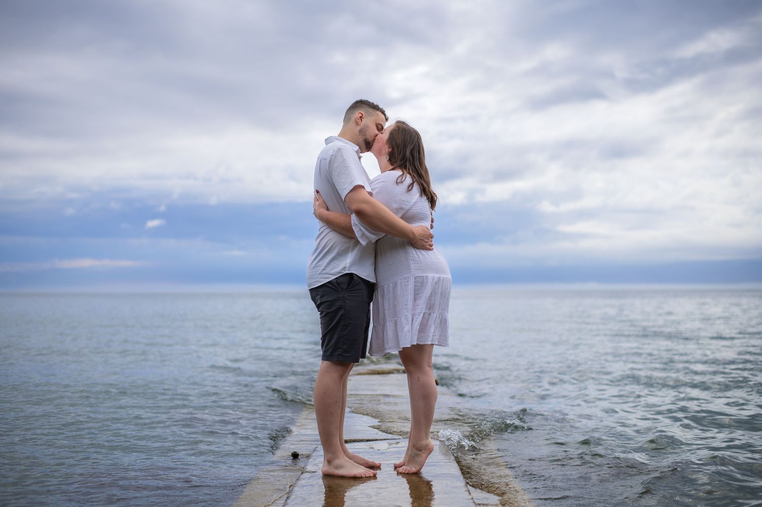 Couple embracing on concrete pier overlooking ocean under cloudy sky.