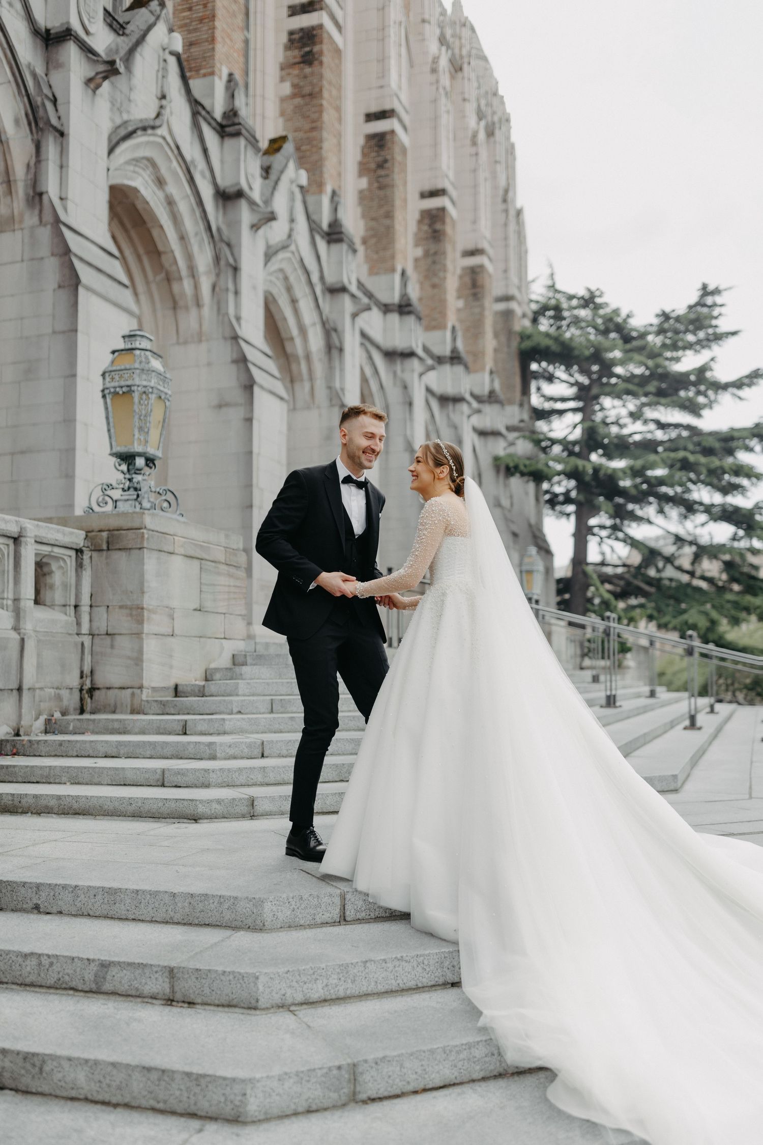 Elegant wedding couple poses on grand stone steps with Gothic architecture backdrop.