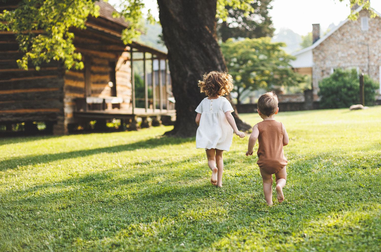 Two children running together in a sun-dappled grassy yard near a rustic cabin.