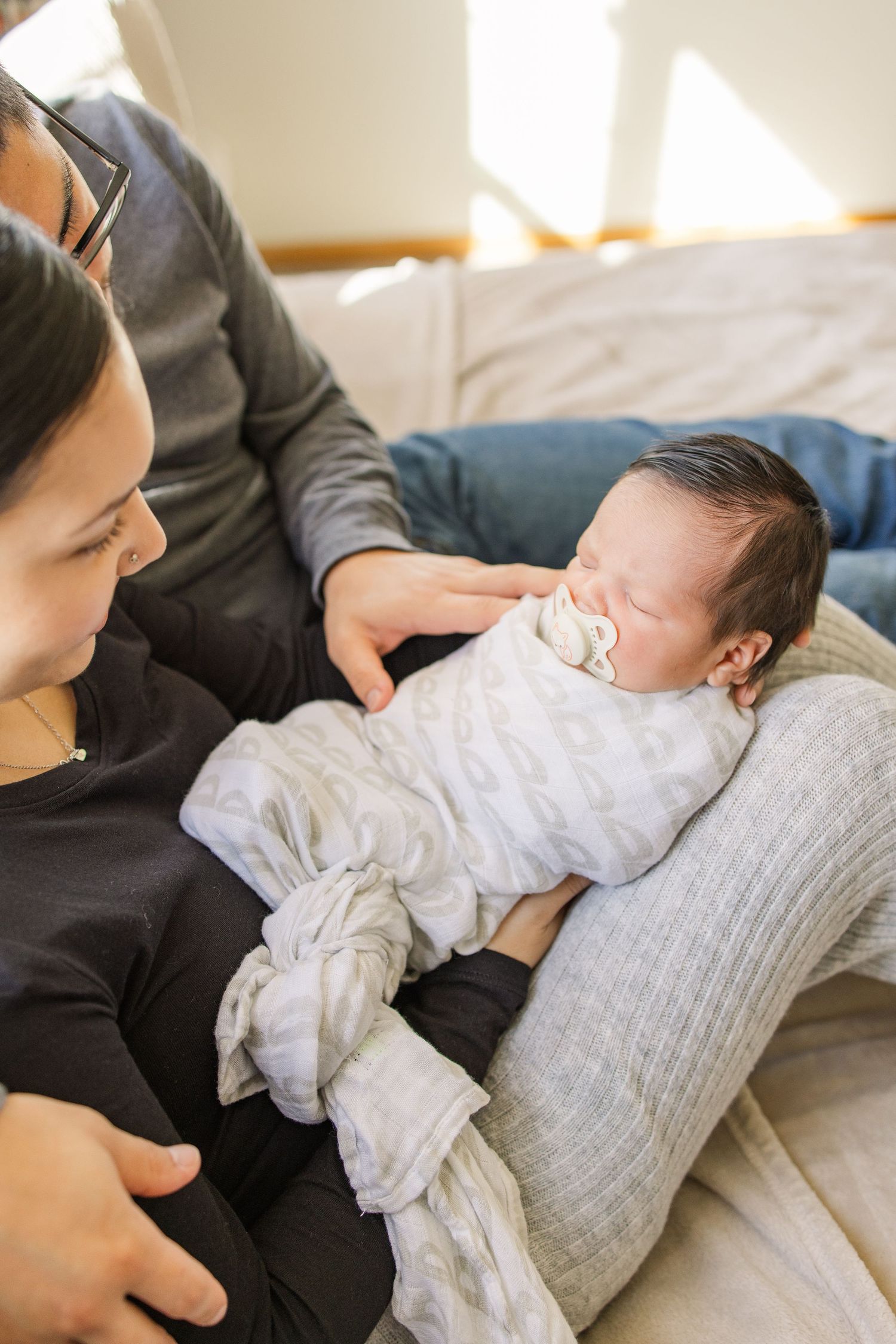 A sleeping newborn in white clothing rests on cushions while being watched.