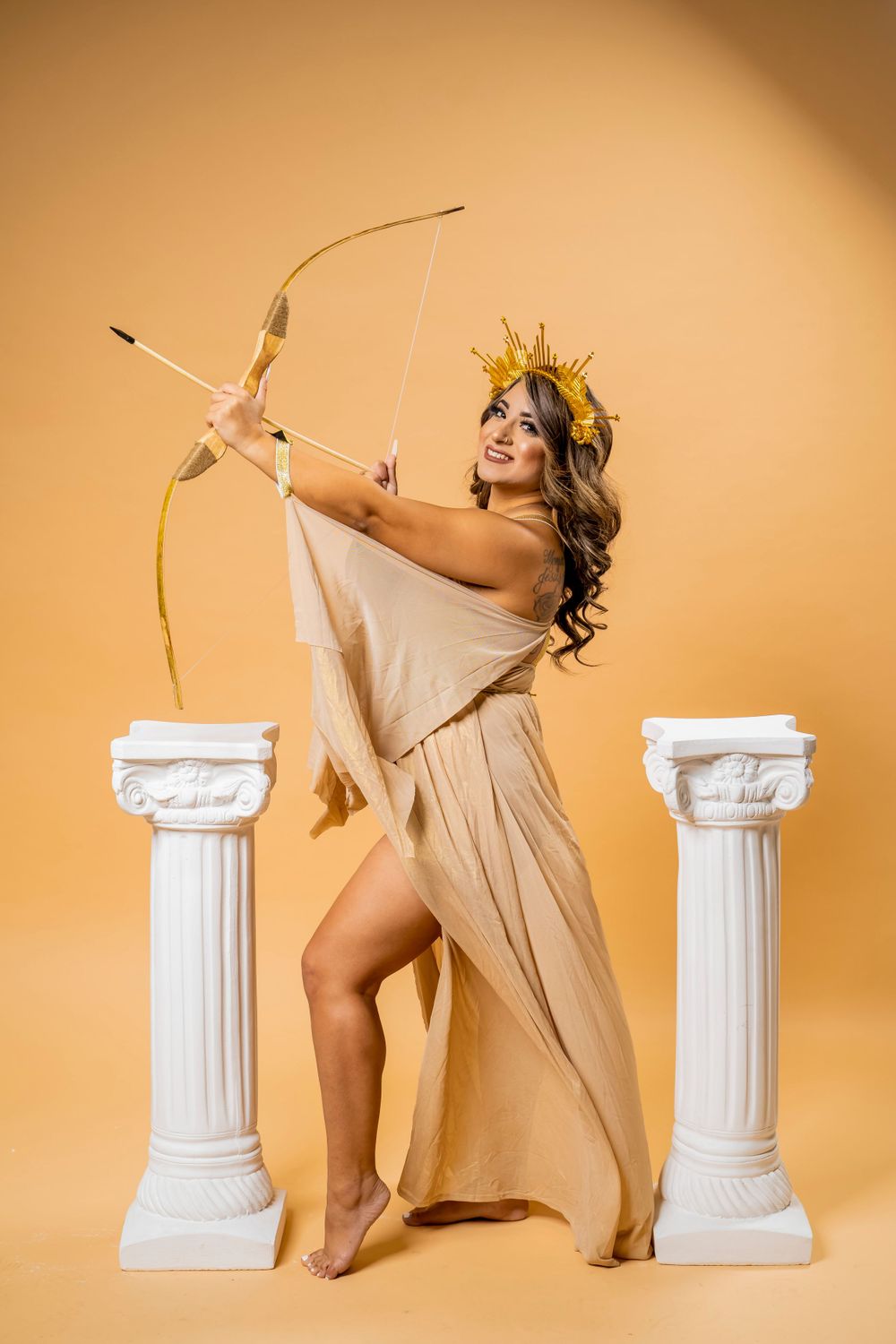 Greek goddess inspired photoshoot featuring bow and arrow with decorative columns against warm beige backdrop.