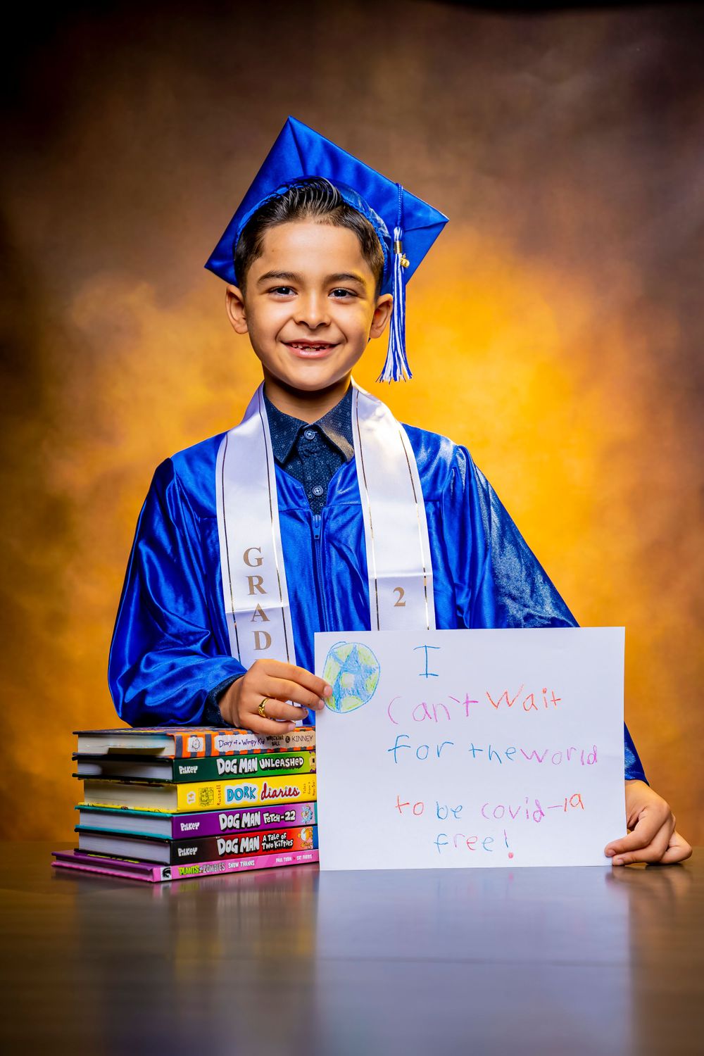 Graduate in blue cap and gown poses with books and handwritten sign.