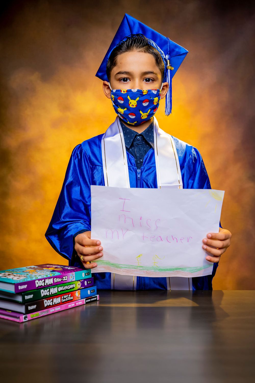 Student in graduation attire wearing face mask holds sign next to stack of books.