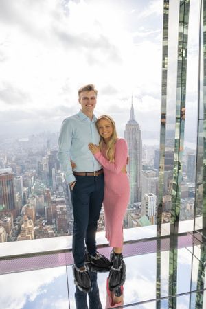 Happy engaged couple posing on glass floor observation deck with Empire State Building and NYC skyline.