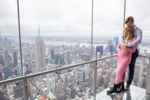 Couple embracing on glass observation deck balcony overlooking the Empire State Building and NYC skyline.