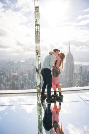 Newly engaged couple sharing a kiss on reflective glass observation deck overlooking New York City skyline.