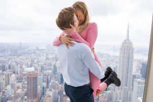 Man lifting woman in celebratory embrace on outdoor observation deck with New York City skyline below.