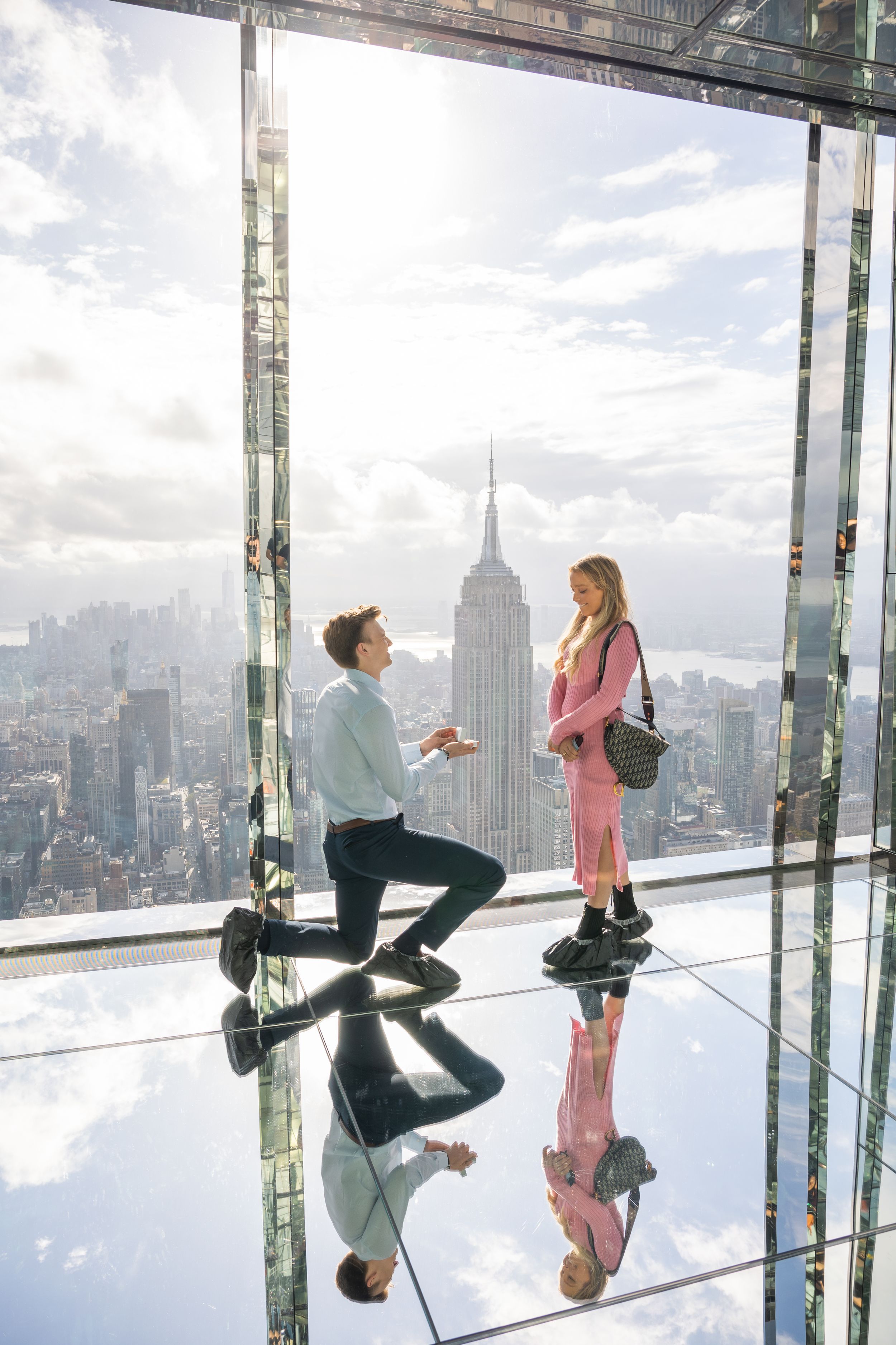 Man proposing on one knee to woman on glass floor observation deck with Empire State Building in background.