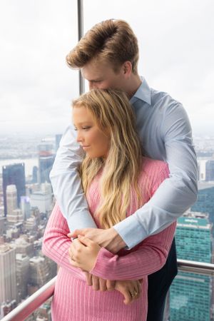 Man tenderly embracing woman from behind on observation deck with New York City skyline in background.